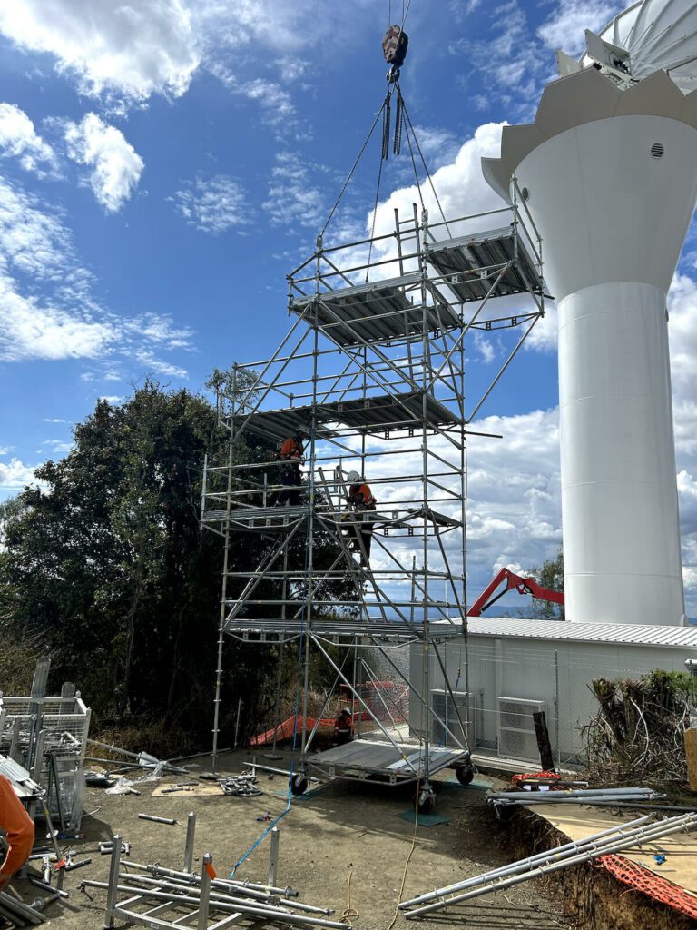 Workers in orange safety gear assemble scaffolding on a construction site under a partly cloudy sky. A crane above lifts a platform to the top of the structure. A large white industrial tower and some trees are visible in the background.