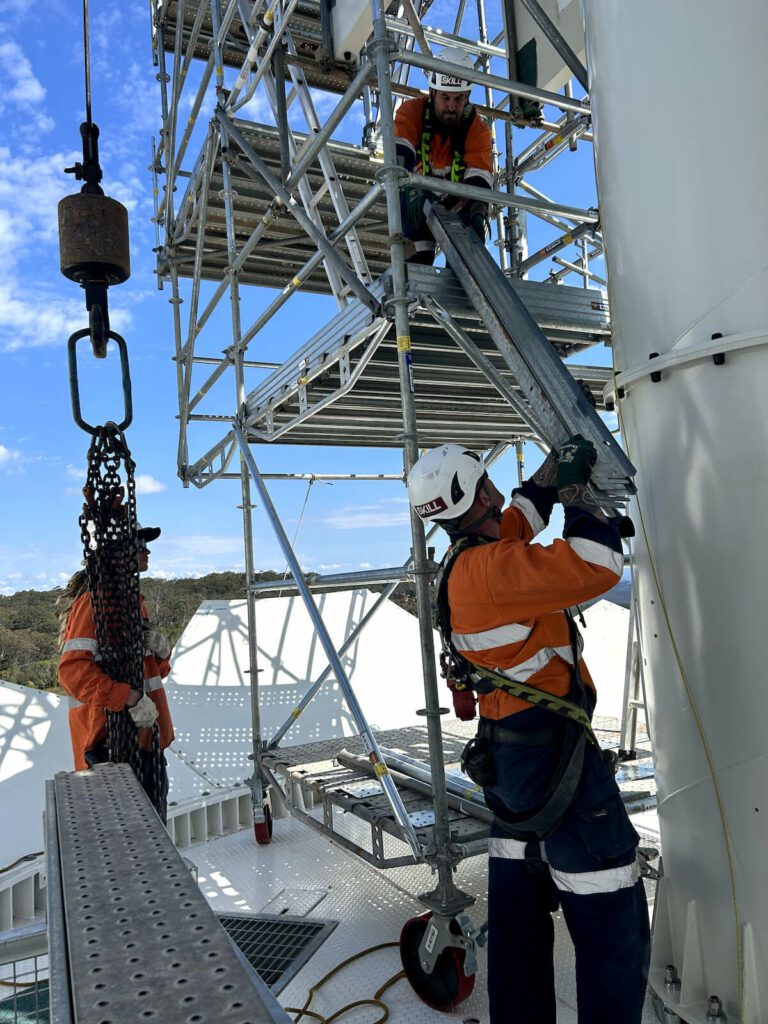 Three construction workers in safety gear, including helmets and harnesses, work on scaffolding attached to a tall structure. One worker holds a large metal panel while another guides it from a higher platform. A third worker monitors the process. A sunny sky is in the background.