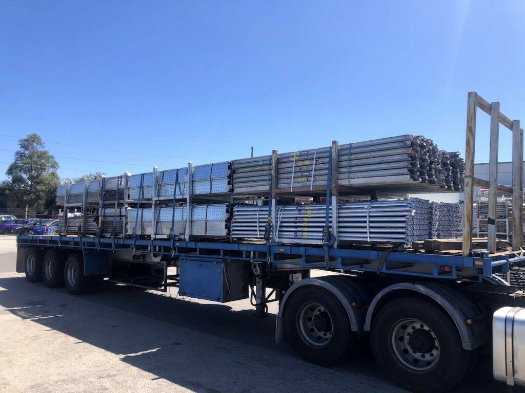 A flatbed truck is parked in an outdoor setting under a clear blue sky, loaded with metal scaffolding pipes and platforms. The cargo appears well-secured with yellow straps and there are other vehicles and trees visible in the background.