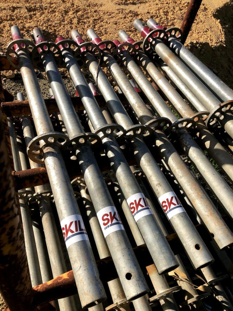 A close-up of a stack of metal scaffolding pipes laid out on the ground. The pipes have a "SKIL" logo on them. The background shows a sandy construction site.