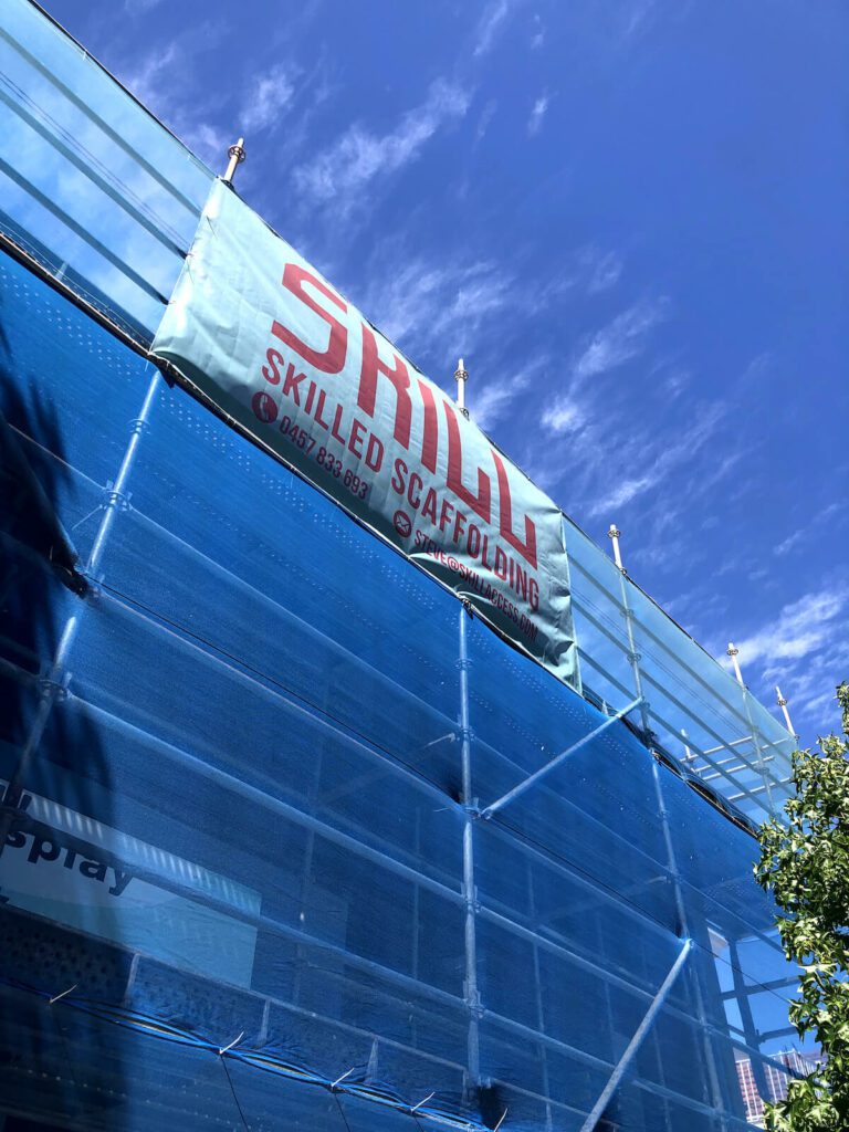 A building under construction with blue scaffolding and netting. A banner attached to the scaffolding reads "Skilled Scaffolding" along with a phone number. The sky is clear and blue with a few wispy clouds. Trees partially frame the right side of the image.
.