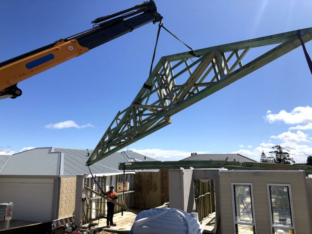 A crane lifts a large wooden roof truss into place at a construction site of a house. Workers on the ground guide the truss. The site is surrounded by completed houses with gray roofs. The sky is clear and sunny, casting sharp shadows.