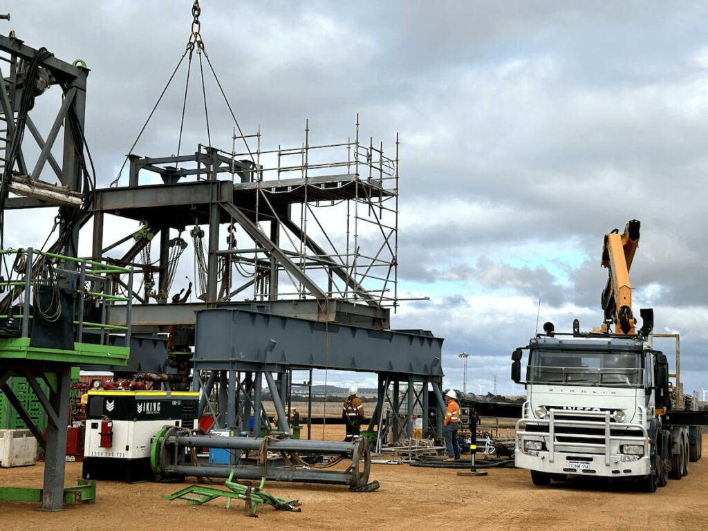 Construction site with workers assembling a large steel structure using a crane. Scaffolding surrounds part of the structure, indicating ongoing work. A white truck with a mounted crane is parked nearby under an overcast sky. Various tools and equipment are scattered around.