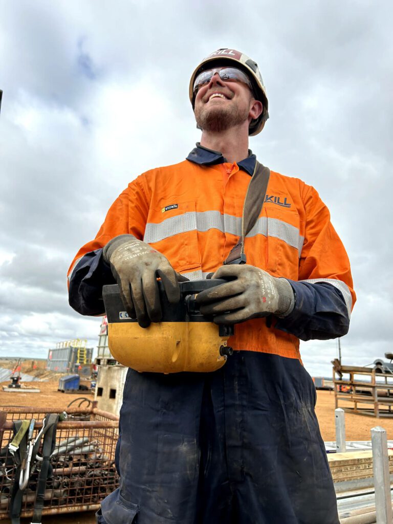 A person in an orange and navy work uniform wearing gloves, sunglasses, and a hard hat stands outdoors, smiling while holding a communication device. Construction equipment and a partly cloudy sky are visible in the background.
