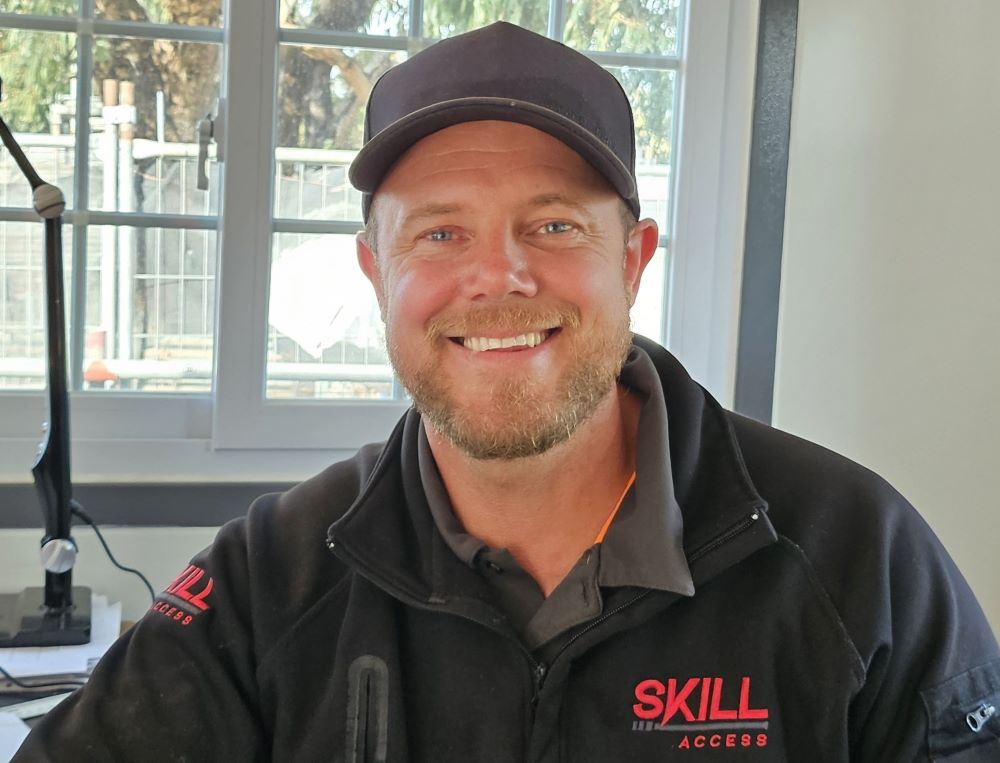 A man wearing a black baseball cap and a black jacket with "SKILL" and "ACCESS" written on it is sitting indoors. He is smiling, and behind him are a window with a view of trees and construction fencing, symbolizing his involvement in access solutions.